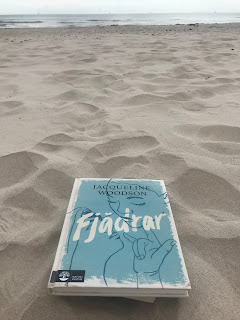 Boken Fjädrar är ljusblå och det syns två ansikten och en hand som gör ett tecken. Boken ligger på en sandstrand med hav i bakgrunden.